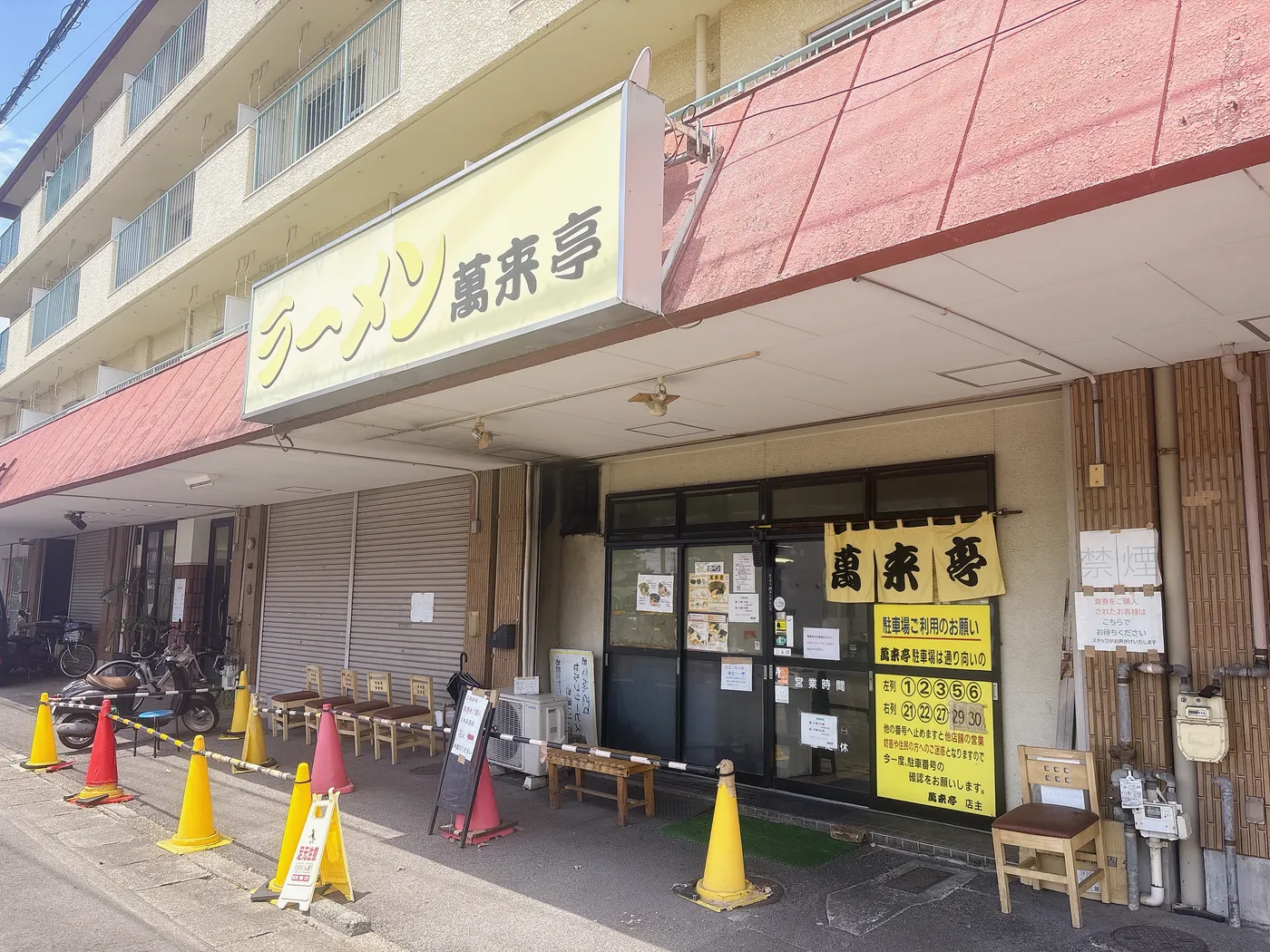 萬来亭の店構え。「ラーメン萬来亭」の看板とのれん、店前に並ぶ待ち椅子