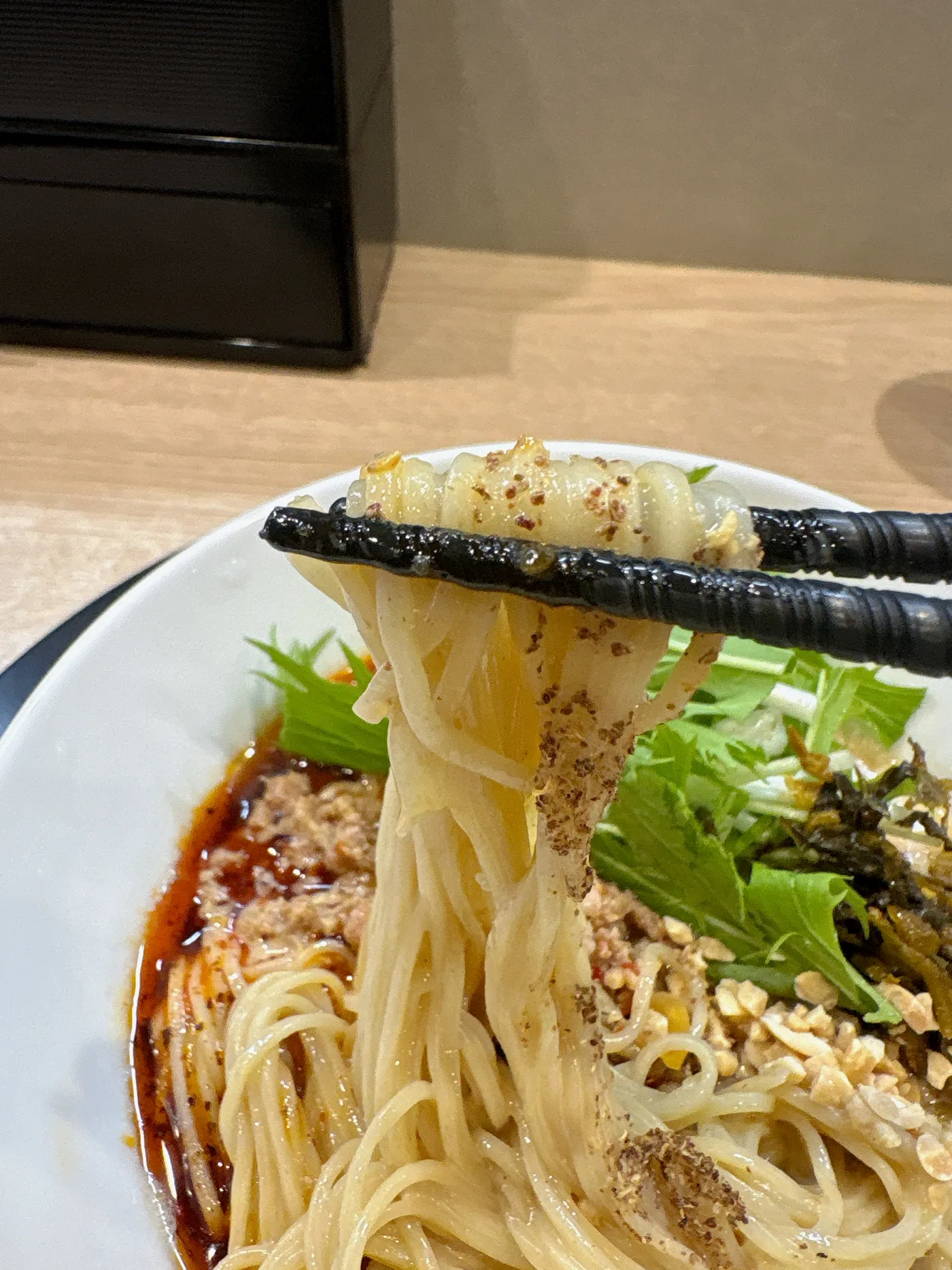 冷やし豆乳担々麺の細めのストレート麺を箸で持ち上げたところ。山椒がまとわりついている